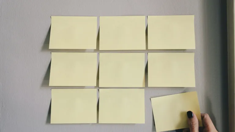 A hand placing the last sticky note into a neat three-by-three grid of notes on a grey wall.